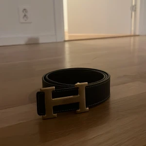 Svart bälte med guldfärgat spänne - Snyggt svart bälte med ett stort guldfärgat H-spänne. Perfekt för att ge en stilren touch till din outfit. Bältet har en klassisk design och passar till många olika stilar.