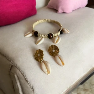 Armband och örhängen med snäckor - Snyggt set med armband och örhängen dekorerade med vita snäckor. Armbandet är knuten med makramé av pappersgarn på en wiretråd, med bruna träpärlor och snäckor. örhängena har en guldig blomformad detalj med hängande snäckor. En vacker kombo till din outfit!