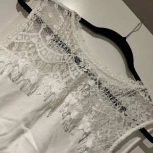 Vit ärmlös topp med spetsdetaljer - Säljer en stilren vit ärmlös topp med vacker spetsdetalj vid halsringningen. Toppen har en v-ringad design.