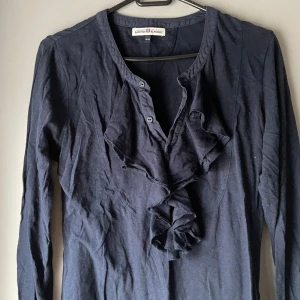 Svart långärmad blus från Hampton Republic - Snygg svart blus från Hampton Republic med volangdetaljer framtill och knappar vid halsen. Perfekt för en stilren look. Långärmad och bekväm passform.