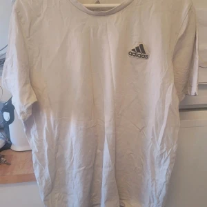 Vit t-shirt från Adidas - Säljer en klassisk vit t-shirt från Adidas med deras ikoniska logga broderad på bröstet. Perfekt för en sportig look eller avslappnad stil. Tillverkad i mjukt material för extra komfort.