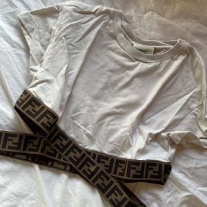 Vit t-shirt från Fendi - Snygg vit t-shirt från Fendi med korta ärmar. Den har ett stilrent och klassiskt utseende med Fendi-logotypen på ett band i brunt och svart. Perfekt för en trendig och avslappnad look.