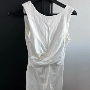 Vit klänning från Crocker Apparel - Elegant vit klänning från Crocker Apparel med en djup v-ringning och dragkedja i ryggen. Klänningen har en stilren design med en draperad detalj framtill som ger en snygg siluett. Perfekt för speciella tillfällen.