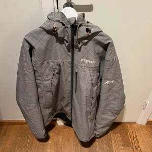 Grå rutig skid jacka från Everest - Säljer en grå rutig skid jacka från Everest med avancerad RECCO-teknologi för lavinräddning. Jackan har en dragkedja framtill och en justerbar huva. Perfekt för kyliga dagar med stil och funktionalitet. Använd få gånger, storlek XL