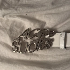 Vitt bälte från Acne Studios - Snyggt vitt bälte från Acne Studios med en iögonfallande silverfärgad logotyp som spänne. Perfekt för att ge din outfit en stilren touch. Bältet har flera hål för justerbar passform.ingen box eller kvitto ingår och väl använt därav priset