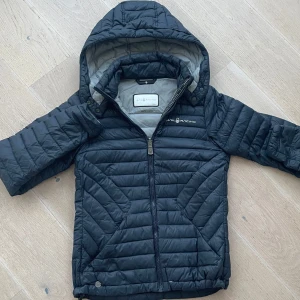 Svart  jacka från Sail Racing - Säljer en svart quiltad jacka från Sail Racing med huva och dragkedja. Jackan har en sportig look och är perfekt för kyligare dagar. Den är designad med Sail Racing-logga på bröstet och har en bekväm passform.