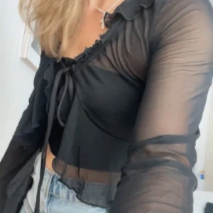 Svart transparent topp / bolero  - Snygg svart transparent blus med volangdetaljer och knytning framtill. Blusen har långa ärmar och en croppad passform, perfekt för en trendig look.