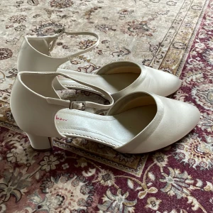 Brudskor  - Eleganta vita pumps med rundad tå och justerbar rem runt ankeln. Skorna har en låg klack och är tillverkade i ett syntetmaterial som ger en stilren look. Perfekta för en formell tillställning eller en kväll ute.