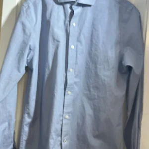 Blå skjorta från Ryan Seacrest Distinction - Säljer en stilren blå skjorta från Ryan Seacrest Distinction. Skjortan har en slim fit passform och är tillverkad i ett non-iron material, perfekt för en smidig och snygg look. Den har långa ärmar och knappar framtill.