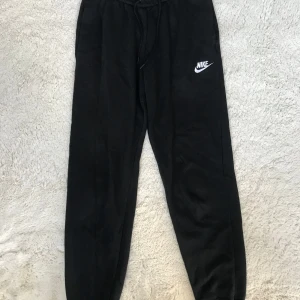 Svarta mjukisbyxor från Nike - Säljer ett par svarta mjukisbyxor från Nike med elastisk midja och dragsko. Byxorna har en avslappnad passform och är perfekta för en bekväm stil. Nike-loggan är tryckt i vitt på benet.
