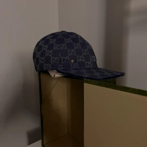 Mörkblå keps från Gucci - Snygg mörkblå keps från Gucci med det ikoniska GG-mönstret. Kepsen har en klassisk design, perfekt för att ge en lyxig touch till din outfit. Den är tillverkad i ett slitstarkt material och har en diskret logotypdetalj i guld. (BETALAR DU HELA PRISET FÅR DU MED GUCCI LÅDAN) vill du sänka priset mer än 500kr blir de utan låda:)