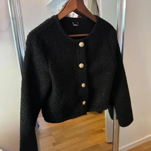 Svart kofta från Gina Tricot - Säljer en snygg svart kofta från Gina Tricot med dekorativa guldknappar framtill. Koftan har en rund halsringning och är tillverkad i ett mjukt material som ger en mysig känsla. Perfekt för kyligare dagar!