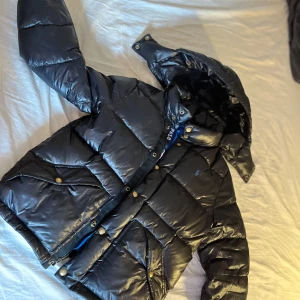 Svart pufferjacka från Polo Ralph Lauren - Säljer en svart pufferjacka från Polo Ralph Lauren. Jackan har en quiltad design med en huva och knappar framtill. Insidan är fodrad med blått tyg för extra värme. Perfekt för kyliga dagar!