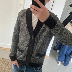 Stickad cardigan - Säljer en stilren grå kofta med svarta detaljer och knappar. Koftan har långa ärmar och ribbade muddar samt fickor framtill. Perfekt för kyligare dagar. Modellen är 186. Skick 8,5/10 ganska ny lite tecken på användning. Skriv för fler frågor eller bilder.