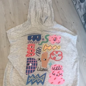 Grå ärmlös hoodie från H&M x Limix - Säljer en unik grå ärmlös hoodie från H&M x Limix med färgglada grafiska tryck på framsidan. Hoodien har en dragkedja och en bekväm huva. Perfekt för en avslappnad stil.