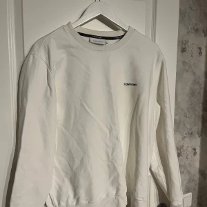 Vit sweatshirt från Calvin Klein - Säljer en stilren vit sweatshirt från Calvin Klein med rund hals och diskret logga på bröstet. Tröjan har långa ärmar och är perfekt för en avslappnad look. Passar bra till både jeans och joggers.