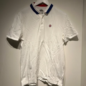 Vit pikétröja från colmar - Snygg vit pikétröja från colmar med blå krage och knappdetaljer. Tröjan har ett litet broderat emblem på bröstet och korta ärmar. Perfekt för en stilren och avslappnad look.