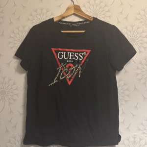 Svart t-shirt från Guess - Snygg svart t-shirt från Guess med det ikoniska triangellogotypen i rött och guld på framsidan. Perfekt för en casual look med en touch av stil. T-shirten har korta ärmar och en rund halsringning.Mycket bra skick som ny.