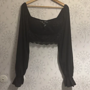 Svart spetsblus med långa ärmar - Snygg svart blus med spetsdetaljer och långa ärmar. Blusen har en croppad design och elastiska ärmslut. Perfekt för en stilren look.