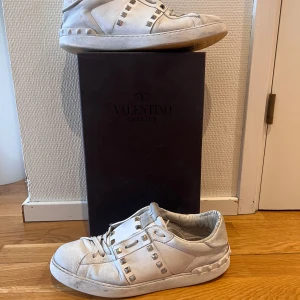 Vita sneakers från Valentino Garavani - Snygga vita sneakers från Valentino Garavani med ikoniska nitar och snörning. Perfekta för en stilren look med en touch av lyx. Skorna har en klassisk design med rund tå och är tillverkade i skinn. De har dock tappat två nitar.