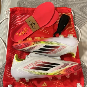 Adidas F50 fotbollsskor - Säljer ett par Adidas F50 fotbollsskor i vitt med svarta och röda detaljer. Skorna har en strömlinjeformad design med snörning och är perfekta för snabba spelare. Innersulor och skohorn ingår.