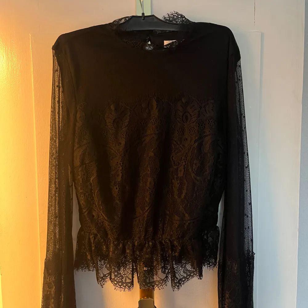 Elegant svart spetsblus från NA-KD med långa ärmar och vackra spetsdetaljer. Blusen har ett prickigt mönster på ärmarna och en fin volangkant nedtill. Perfekt för en stilfull look.. Puserot.