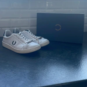 Vita sneakers från Fred Perry - Snygga vita sneakers från Fred Perry i skinn med klassisk logga på sidan. Skorna är i storlek 42 och nypriset ligger på 1100kr, skick 9/10. Boxen följer självklart med. Skorna har snörning och en stilren design som passar perfekt till vardags. De har en mörk detalj vid hälen som ger en extra touch. Otroligt stilrena och perfekta skor för att göra din stil ännu coolare och för en causal look 👀mega bra pris just nu på dessa fina skorna!! Tveka inte på att höra av er ✅