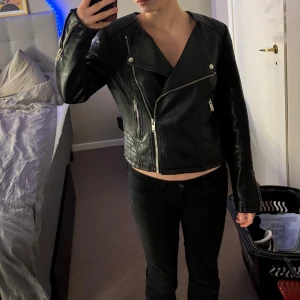 Svart skinnjacka med dragkedja - Snygg svart skinnjacka med asymmetrisk dragkedja och coola detaljer. Jackan har långa ärmar och en klassisk bikerstil. Perfekt för att ge din outfit en edgy touch.