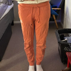 Orange mjukisbyxor från Nike - Säljer ett par bekväma orange mjukisbyxor från Nike. Byxorna har en elastisk midja med snörning och vita detaljer längs sidorna. Perfekta för en avslappnad stil.
