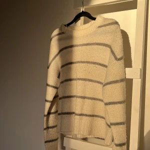 Vit och grå randig stickad tröja - Mysig vit stickad tröja med grå ränder. Perfekt för kyliga dagar med sin mjuka och varma känsla. Tröjan har långa ärmar och en klassisk rund halsringning. Ett måste för garderoben!