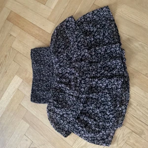 Svart blommig kjol - Säljer en söt svart kortkjol med små vita blommor. Kjolen har en volangdetalj och en elastisk midja för bekväm passform. Perfekt för sommarens varma dagar.