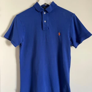Blå pikétröja från Ralph Lauren - Säljer en snygg blå pikétröja från Ralph Lauren i storlek S. Tröjan har en krage med två knappar och den broderade logotypen i orange på bröstet. Perfekt för en stilren och avslappnad look!