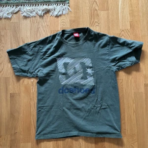Grön t-shirt från DC Shoes - Säljer en grön t-shirt från DC Shoes i storlek Large. Den har ett stort tryck med DC-loggan och texten 'DCSHOECOUSA' på baksidan. Materialet känns mjukt och bekvämt.