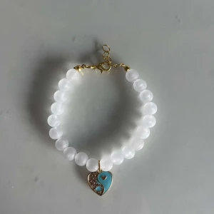 Vitt pärlarmband med hjärthänge - Snyggt armband med vita pärlor och ett unikt hänge i form av ett hjärta. Hänget är delvis turkost och delvis guldigt med små hjärtan inuti. Perfekt för att ge en romantisk touch till din outfit!