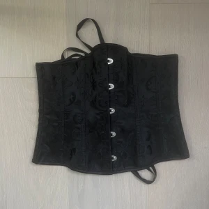 Svart korsett med snörning - Snygg svart korsett med dekorativa knappar framtill och snörning bak. Perfekt för en festlig kväll eller en speciell tillställning. Den har en elegant design med ett subtilt mönster och justerbara band för bästa passform.