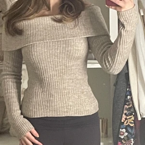 Beige offshoulder stickad tröja - Säljer en snygg beige offshoulder stickad tröja med ribbad struktur. Tröjan har långa ärmar och en bred krage som ger en stilren look. Perfekt för kyligare dagar!