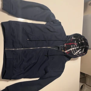 Mörkblå hoodie från Burberry - Säljer en snygg mörkblå hoodie från Burberry.                                           med dragkedja och klassiskt rutigt mönster i luvan. Perfekt för en stilren look.                                   Hoodien har långa ärmar och praktiska fickor framtill.                                  Fick i present så vet ej om den är äkta men riktigt fin kvalitet 