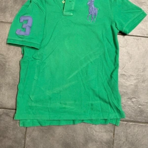 Grön pikétröja från Ralph Lauren - Säljer en grön pikétröja från Ralph Lauren med korta ärmar och en stor lila logga på bröstet. Tröjan har även en lila siffra '3' på ärmen. Perfekt för en sportig look!