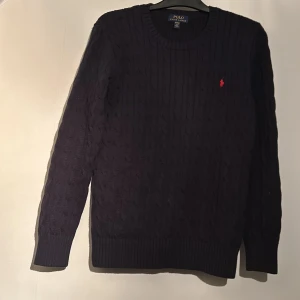 Mörkblå stickad tröja från Ralph Lauren - Säljer en stilren mörkblå stickad tröja från Ralph Lauren med det klassiska röda logotypbroderiet på bröstet. Tröjan har långa ärmar och ribbstickade muddar vid ärmslut och nederkant. Perfekt för kyligare dagar. KOM MED BUD!!!