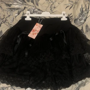 Svart kjol med spets och volang - Snygg svart kjol med flera lager av spets och volangdetaljer. Kjolen har en elegant och lekfull design med satinband som ger en extra touch. Perfekt för att skapa en unik stil. Aldrig använt. 
