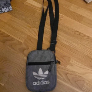 Grå axelväska från Adidas - Snygg grå axelväska från Adidas med justerbar rem och dragkedja. Väskan har det klassiska Adidas-loggan framtill och text på baksidan. Perfekt för att bära med sig det nödvändigaste på ett stilrent sätt.