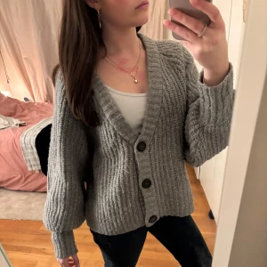 Grå stickad kofta från Stories - Mysig grå stickad kofta från Stories med stora knappar framtill. Perfekt för kyliga dagar med sin ribbade design och långa ärmar. Passar bra över en t-shirt eller linne för en avslappnad look.