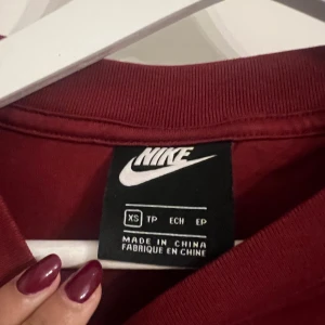 Vinröd croppad tröja från Nike - Säljer en vinröd croppad tröja från Nike med långa ärmar. Tröjan har en stilren design med Nike-loggan tryckt på ena ärmen och ryggen. Perfekt för en sportig look eller avslappnad stil. Passar bra till jeans eller träningsbyxor.