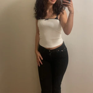 Vit ribbad linnetopp med spetsdetaljer - Säljer en söt vit ribbad linnetopp med smala axelband och svart spetskant. Toppen har en liten svart rosett framtill som ger en extra touch. Perfekt för varma dagar eller som ett lager under en kofta.