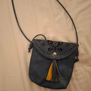 Grå/blå axelväska med fjärilsmönster - Snygg grå/blå axelväska i äkta läder med ett utskuret fjärilsmönster på locket. Väskan har en dekorativ tofs i gult och stängs med en knapp. Perfekt för att ge en stilfull touch till din outfit.