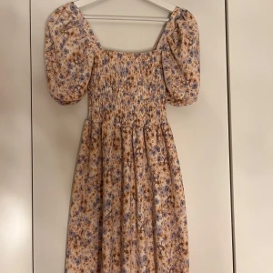 Blommig sommarklänning med puffärmar - Charmig kort blommig klänning med puffärmar och ribbad överdel för bättre passform. Klänningen har en ljus rosa bas med blommönster i lila och beige toner. Perfekt för en somrig look.