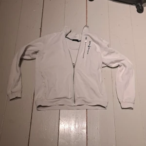 Vit hoodie från Sail Racing - Säljer en vit hoodie från Sail Racing med dragkedja framtill. Tröjan har långa ärmar och en hög krage. Perfekt för kyliga dagar. Hör av dig vid intresse! Vi har katt och hund hemma och köparen står för frakten. Den är inte mycket använd men lågt pris pga fläckarna.