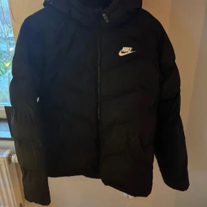 Svart dunjacka från Nike modell - Säljer en svart dunjacka från Nike med dragkedja och huva. Jackan har en klassisk pufferdesign och är perfekt för kyligare väder. Den har långa ärmar och en diskret Nike-logga på bröstet.