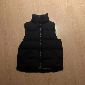 Svart dunväst från Boomerang - Säljer en svart dunväst från Boomerang med dragkedja och broderad logotyp. Perfekt för kyligare dagar och enkel att matcha med andra plagg. Västen har en stilren design och hög krage för extra värme.