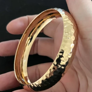 Guldfärgat armband med hamrad yta - Elegant guldfärgat armband med en stilren hamrad yta som ger en unik textur. Perfekt för att lägga till en touch av glamour till din look. Armbandet har en rund form och är lätt att matcha med andra smycken.